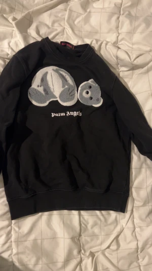 Svart Palm Angels sweatshirt med björn - Svart sweatshirt från Palm Angels med stort broderat motiv av en nalle på bröstet och vit logotyp under. Tröjan har rund hals och långa ärmar, perfekt för dig som gillar streetwear och coola detaljer. Mycket bra skick använd fåtalgånger därför för endast 300kr