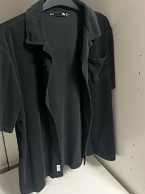 Svart frottéskjorta från 157 - Svart skjorta från 157 i mjukt frottématerial, med korta ärmar och klassisk krage. Skjortan har en bröstficka och knäppning framtill. Perfekt för dig som gillar chill och enkel stil.