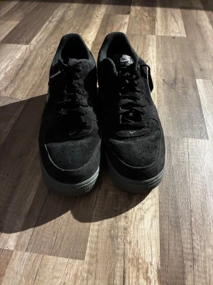 Svarta Nike Air Force 1 sneakers - Säljer ett par svarta Nike Air Force 1 sneakers med grå sula och klassisk swoosh på sidan. Skorna har snörning och är tillverkade i mocka med perforerad tå för extra ventilation. Perfekt för dig som gillar stilrena och tidlösa sneakers. Använda en gång 