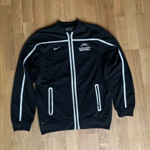 Svart Nike Dri-Fit basketjacka - Svart Nike Dri-Fit jacka med vita ränder och dragkedja framtill. Broderad Floyd County Basketball-logga på bröstet och klassisk Nike Swoosh. Jackan har ribbade muddar och två fickor med vita detaljer. Perfekt för basketträning eller chill. Armländ: 62cm. Längd 64cm. Pris kan diskuteras