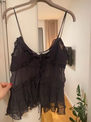 Svart volang linnetopp från Zara - Säljer en svart, tunn och luftig linnetopp från Zara med volangdetaljer och snörning framtill. Toppen har smala axelband och är tillverkad i ett lätt, transparent material som ger en cool och avslappnad vibe. Perfekt för varma dagar eller att styla lager på lager.