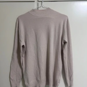 Beige polotröja med ribbad hals - Snygg beige polotröja med ribbad hals och muddar vid ärmslut och nederkant. Tröjan har en enkel och stilren design med lång ärm, perfekt för lager-på-lager under kyliga dagar. Materialet känns mjukt och lätt mot huden.