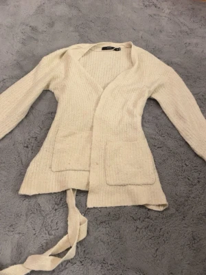 Beige stickad kofta med knytband - Mysig beige stickad kofta från Vera moda med långa ärmar, två stora fickor framtill och ett knytband i midjan. Perfekt för lager-på-lager och chill dagar. Koftan har en öppen design och ribbad struktur som ger en avslappnad vibe. Kan eventuellt tänka mig gå ner i pris