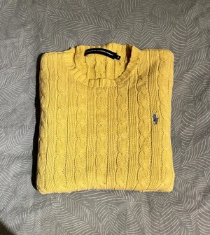 Gul kabelstickad tröja från Ralph Lauren - Säljer en gul kabelstickad tröja från Ralph Lauren Sport i storlek S. Tröjan har rund hals, långa ärmar och ett blått broderat logotyp på bröstet. Materialet är mjuk bomull och den har ribbade muddar vid ärmslut och nederkant. Perfekt för dig som gillar färg och klassisk stil.