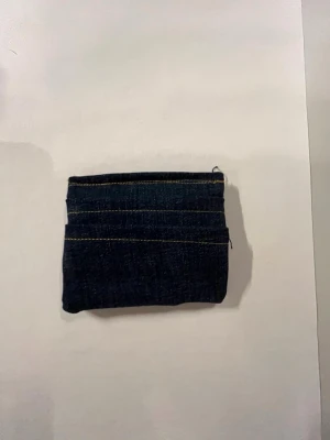 Mörkblå jeansplånbok med sömmar - Säljer en liten och smidig plånbok i mörkblått jeanstyg med synliga kontrastsömmar. Perfekt för kort och småmynt, enkel design med flera fack. Plånboken har en rektangulär form och är gjord av denimtyg.