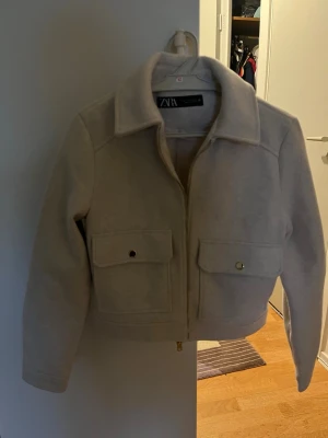 Beige bomberjacka från Zara - Snygg beige bomberjacka från Zara med klassisk krage och två stora fickor framtill med knappstängning. Jackan har dragkedja och är tillverkad i ett mjukt material som känns skönt mot huden. Perfekt för dig som gillar stilrena och enkla plagg.