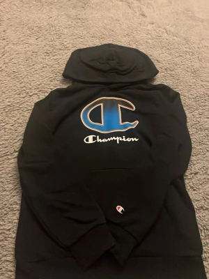 Champion hoodie för barn  - Det är en svart hoodie från Champion. 	•	På bröstet sitter den stora klassiska Champion-loggan – ett stort blått “C” med vit/orange kant 	•	Under loggan står “Champion” i vitt 	•	Tröjan har huva och känguruficka framtill 	•	På ärmen syns den lilla Champion-patchen 	•	Ser ut att vara i tjock, mjuk sweatshirt-kvalitet, ganska clean och sportig sti