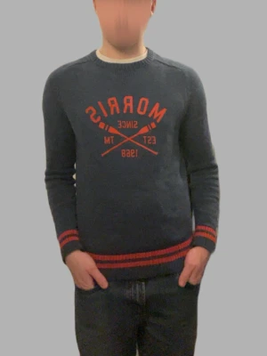 Mörkblå stickad tröja Morris Gents - Snygg mörkblå stickad tröja från Morris Gents i storlek small. Tröjan har röd text och tryck framtill samt röda ränder vid ärmslut och nederkant. Rund hals och långärmad modell, perfekt för dig som gillar klassisk stil med en sportig touch.