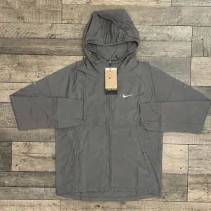 Grå vindjacka från Nike med huva - Säljer en grå vindjacka från Nike med huva och hel dragkedja framtill. Jackan har två sidofickor och en stilren design med Nike-logga på bröstet. Perfekt för dig som gillar sportig och clean stil. Materialet är lätt och känns skönt mot kroppen.