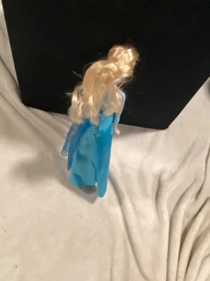 Blå glittrig väst till docka - Säljer en blå glittrig väst till docka, med lång transparent cape baktill. Västen har en hjärtformad överdel och är gjord i syntetmaterial med glittriga detaljer framtill. Perfekt för att ge dockan en magisk look.