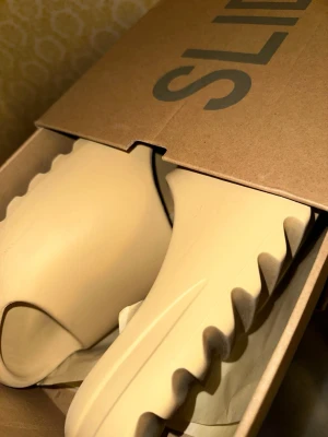 Adidas Yeezy Slide beige sandaler - Adidas Yeezy Slide i beige med chunky, räfflad sula och öppen tå. Slidesen har en minimalistisk design och är tillverkade i syntetmaterial som ger en mjuk och bekväm känsla. Perfekt för chill dagar och sommarhäng. Ikonisk Yeezy-look med Adidas-logga i fotbädden.