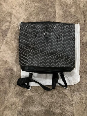 Svart ryggsäck  - Säljer en svart ryggsäck från  med ikoniskt grafiskt mönster i vitt. Väskan har detaljer i skinn, justerbara axelband och metallspännen. Perfekt för dig som vill ha en stilren och lyxig accessoar med plats för allt du behöver.