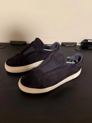 Mörkblå mockasneakers från ARIGATO - Snygga mörkblå sneakers från ARIGATO i mocka med vit platt sula. Skorna har en minimalistisk design utan snörning och elastiska paneler på ovansidan för enkel påtagning. Perfekta för dig som gillar stilrena och bekväma skor med premiumkänsla.