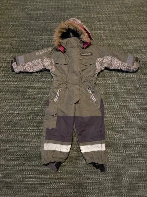 Grå Didriksons barnoverall - Säljer en grå overall från Didriksons med reflexdetaljer, avtagbar huva med fuskpäls och mönstrade partier på ärmar och huva. Overallen har dragkedja framtill, förstärkta knän och elastiska benslut med fotremmar. Perfekt för kalla dagar utomhus.