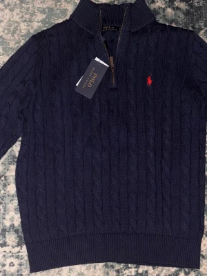 Marinblå stickad tröja Polo Ralph Lauren - Säljer en marinblå stickad tröja från Polo Ralph Lauren med kabelstickat mönster och halv dragkedja. Tröjan har lång ärm, ribbade muddar och den klassiska röda loggan broderad på bröstet. Perfekt för kyliga dagar!