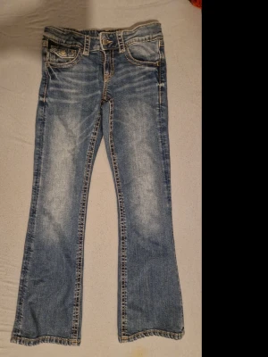 Blå bootcut jeans Gina Tricot 152 - Säljer ett par blå bootcut jeans från Gina Tricot i storlek 152. Jeansen har klassisk femficksmodell, snygga slitningar framtill och kontrastsömmar. Materialet är jeans med en skön passform och bakfickor med knappdetaljer.