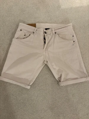 Dondup Beige shorts i bomull, storlek 31 - Säljer ett par ljusbeige shorts i bomull med klassisk femficksdesign och uppvikta benslut. De har knappgylf och detaljer i metall. Perfekta för sommaren och riktigt sköna att bära till t-shirt eller skjorta.