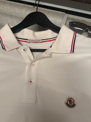 Vit Moncler pikétröja med rödblå detaljer - Säljer en klassisk vit pikétröja från Moncler med röd och marinblå rand på krage och ärmslut. Tröjan har knappslå framtill och Moncler-logga broderad på bröstet. Skjortan skulle behövas strykas och det gör jag innan jag fraktar den😁 Tveka inte att höra av dig om du undrar något eller vill se fler bilder!
