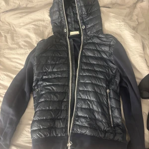 Moncler cardigan  - Snygg svart dunväst från Moncler med huva och dubbla dragkedjor framtill. Västen har quiltad design och två fickor med dragkedja. Materialet är glansig polyester och dun, perfekt för lager på lager under höst och vinter.