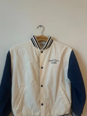 Vit och marinblå bomberjacka från Zara - Snygg bomberjacka från Zara i vitt med marinblå ärmar och detaljer. Jackan har ribbade muddar med ränder, tryckknappar framtill och broderad text 'Travel Fever' på bröstet och ryggen. Tillverkad i polyester och har två fickor fram.