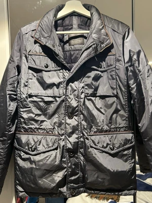 Svart/gråa  pufferjacka från Massimo Dutti - Snygg svart pufferjacka från Massimo Dutti med fyra stora fickor framtill och knappar. Jackan har hög krage med diskret kontrastkant och är tillverkad i glansig polyester. Perfekt för kyligare dagar och har en klassisk, stilren look.