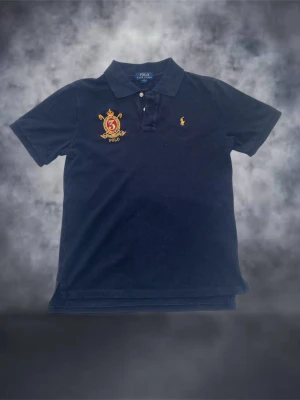 Marinblå pikétröja från Ralph Lauren - Säljer en marinblå pikétröja från Ralph Lauren med klassisk krage och knappar. Tröjan har ett broderat gult polospelare-märke på bröstet och en stor broderad patch med nummer 3 och detaljer i rött, blått och guld. Tillverkad i bomull för en skön känsla.