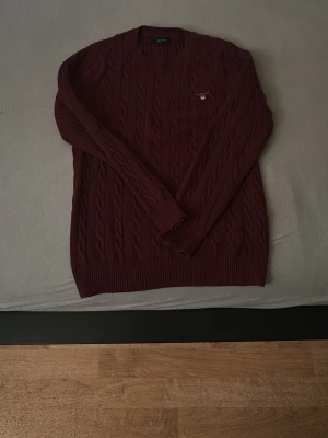 Vinröd kabelstickad tröja från GANT - Säljer en vinröd kabelstickad tröja från GANT med rund halsringning och diskret logga på bröstet. Tröjan har långa ärmar och ribbade muddar vid ärmslut och nederkant. Perfekt för höst och vinter när du vill vara både snygg och varm.