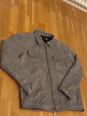 Grå mocka overshirt med dragkedja - Säljer en stilren grå overshirt i mocka-look med raka linjer och två stora bröstfickor. Jackan har en klassisk krage, långa ärmar med knappdetaljer vid mudden och hel dragkedja framtill. Perfekt för dig som gillar minimalistisk och clean stil.