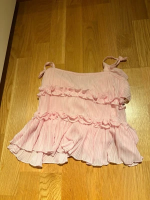 Rosa volangtop från Amisu XS - Supersöt rosa topp från Amisu i storlek XS. Toppen har tunna axelband med knyt, volanger och ribbad struktur. Materialet är lätt och luftigt, perfekt för varma dagar. Snygg detalj med knyt framtill och flera lager volanger som ger en romantisk vibe.
