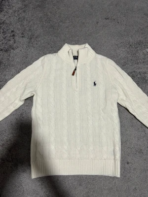 Vit kabelstickad tröja Polo Ralph Lauren - Vit kabelstickad tröja från Polo Ralph Lauren med halv dragkedja och ribbade muddar. Klassisk logga broderad på bröstet och hög krage. Perfekt för lager på lager under höst och vinter. Har använt den bara några gånger. Priset kan diskuteras!