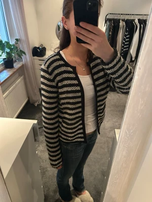 Randig svartvit stickad kofta - Säljer en stickad kofta med svart och vita ränder. Koftan har rund halsringning, lång ärm och är öppen framtill med två ”krok” knappar. Materialet är grovstickat och en ficka finns på vadera sida