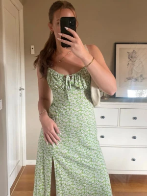 Grön blommig klänning med slits - Superfin grön klänning med vita blommor, smala axelband och söt snörning framtill. Klänningen har en slits framtill och är perfekt för varma dagar. Materialet känns lätt och luftigt, och modellen är figurnära men ändå bekväm.