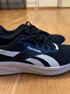 Svarta och vita Reebok sneakers - Säljer ett par snygga Reebok sneakers i svart och vitt med klassisk logga på sidan och hälen. Skorna har snörning, rund tå och vit platt sula. Ovandelen är i textil och syntet, vilket gör dem lätta och bekväma för vardagsbruk eller träning.