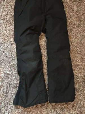Svarta täckbyxor med vida ben - Säljer ett par svarta täckbyxor med bred passform och raka ben. Perfekta för kalla dagar och vinteraktiviteter. Byxorna har praktiska fickor och är tillverkade i ett slitstarkt syntetmaterial som står emot vind och kyla.