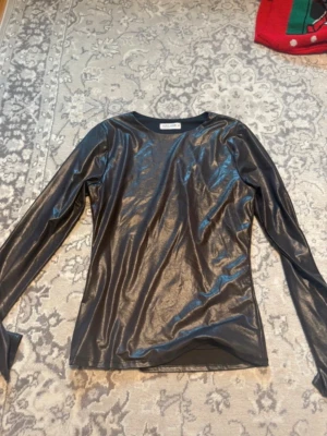 Svart glansig långärmad topp Popular 21 - Säljer en svart långärmad topp från Popular 21 i storlek M. Toppen har en glansig finish som ger en cool look och är tillverkad i ett syntetmaterial som känns mjukt mot huden. Passformen är normal och toppen har en enkel, stilren design utan extra detaljer.