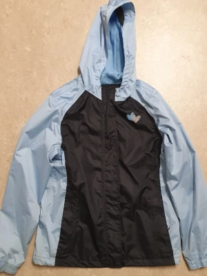 Vindjacka - Snygg vindjacka från Crivit i ljusblått och svart med huva och dragkedja. Jackan har praktiska kardborrestängningar, sidfickor och meshfoder på insidan. Perfekt för blåsiga dagar och enkel att matcha med din vardagsstil. Har en fläck.