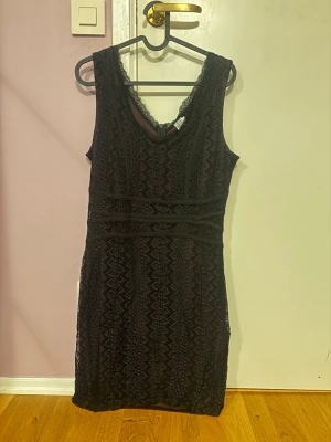 Svart spetsklänning från Saint Tropez - Säljer en svart kortklänning från Saint Tropez med snygg spets och v-ringning. Klänningen är ärmlös och har ett fint mönster i spetsen samt dragkedja i sidan. Perfekt för dig som vill ha en elegant look med coola detaljer.