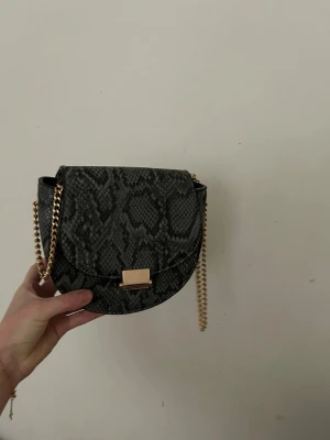 Svart/grå axelväska med ormmönster - Säljer en svart axelväska med snyggt ormmönster och guldfärgad kedja. Väskan har rundad form, guldfärgat spänne och ett praktiskt innerfack med dragkedja. Perfekt storlek för det viktigaste och ger en cool touch till din outfit. Aldrig använd. 
