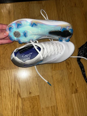 Vita Skechers Hyper Burst Pro fotbollsskor - Säljer ett par vita Skechers Hyper Burst Pro fotbollsskor med blå detaljer och cool iriserande sula. Skorna har snörning, låg profil och är tillverkade i syntetmaterial för lätt känsla på planen. Perfekta för dig som vill sticka ut med stilrena och moderna dobbar. Nypris är 2599!