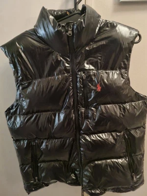 Svart dunväst från Polo Ralph Lauren - Snygg svart dunväst från Polo Ralph Lauren med glansig finish och hög krage. Västen har dragkedja framtill, tre praktiska fickor med dragkedja och den klassiska röda loggan broderad på bröstet. Perfekt för lager på lager under kyliga dagar.