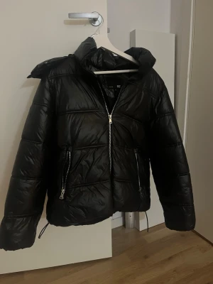 Svart pufferjacka - Säljer en svart pufferjacka från 157 med hög krage och huva. Jackan har två stora fickor med dragkedjor och justerbar dragsko nertill. Perfekt för kalla dagar och riktigt skön att bära. Passformen är normal och materialet är glansigt syntet.