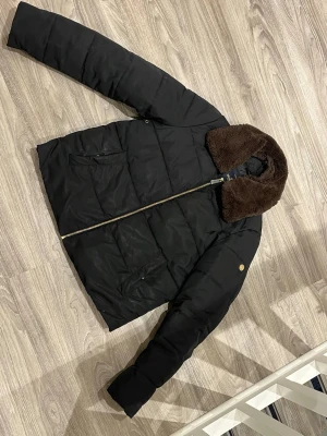 Svart pufferjacka med brun krage - Säljer en svart pufferjacka med en stor, mjuk brun fuskpäls-krage. Jackan har guldfärgad dragkedja, två fickor framtill och quiltad design. Perfekt för kalla dagar och riktigt snygg till jeans eller cargobyxor.