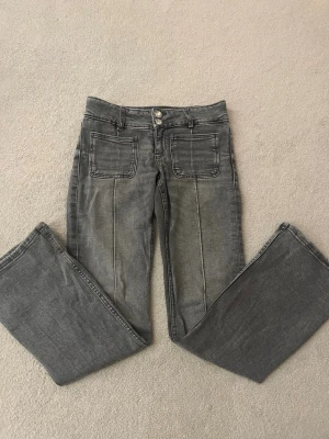 Grå bootcut jeans med fickor - Snygga grå jeans med bootcut-modell och raka ben. Byxorna har två stora framfickor och markerade sömmar längs benen. Stängs med dubbla knappar och dragkedja. Perfekta för en chill och trendig look.