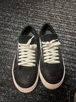 Svarta sneakers från Rick Owens - Säljer ett par svarta sneakers från Rick Owens med vit sula och vita snören. Skorna har en kontrasterande vit detalj på sidan och är tillverkade i mocka och skinn. Klassisk låg modell med rund tå och platt sula, perfekta för en clean och edgy stil. Pris kan diskuteras 👌