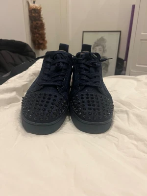 Mörkblå loubs - Säljer ett par mörkblå sneakers med coola svarta nitar på tån. Skorna har snörning och är gjorda i mocka med en platt sula. Perfekta för dig som vill sticka ut med en edgy stil och gillar detaljer som syns. Pris kan diskuteras vid snabb affär