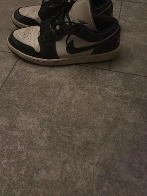 Nike Air Jordan 1 Low svart/vit US11 - Säljer ett par Nike Air Jordan 1 Low i svart och vitt. Klassisk siluett med svart swoosh, perforerad tåbox och låga kanter. Skorna har snörning och rund tå. Tillverkade i skinn och syntetmaterial. Perfekta för dig som gillar streetwear och sneakers med ikonisk design.