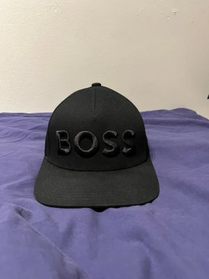 Svart keps från BOSS - Säljer en stilren svart keps från BOSS med broderad logga framtill. Kepsen har böjd skärm, justerbart spänne bak och är tillverkad i bomull. Perfekt accessoar för en clean och modern look.