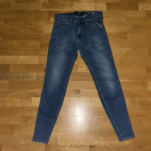Replay Anbass blå jeans, storlek 29/32 - Snygga blå Replay Anbass jeans med klassisk femficksdesign och smal passform. Jeansen har en mörk tvätt med lätt slitningar och ett litet hål  och är tillverkade i stretchigt denim för extra komfort. Perfekt för dig som gillar en modern och stilren look.