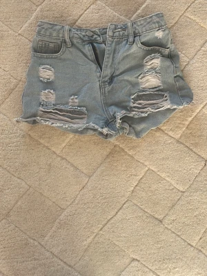 Ljusa blå jeansshorts med slitningar - Säljer ett par ljusblå jeansshorts med råa kanter och stora slitningar framtill. Klassisk femficksmodell med bälteshällor och knappgylf. Perfekta för sommaren och ger en avslappnad vibe.