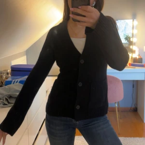 Svart stickad kofta från Wera - Mysig svart stickad kofta från Wera med v-ringning och stora knappar framtill. Koftan har två fickor och långa ärmar med breda ribbade muddar. Perfekt att slänga på sig över en t-shirt eller linne för en chill look. Nypris 899